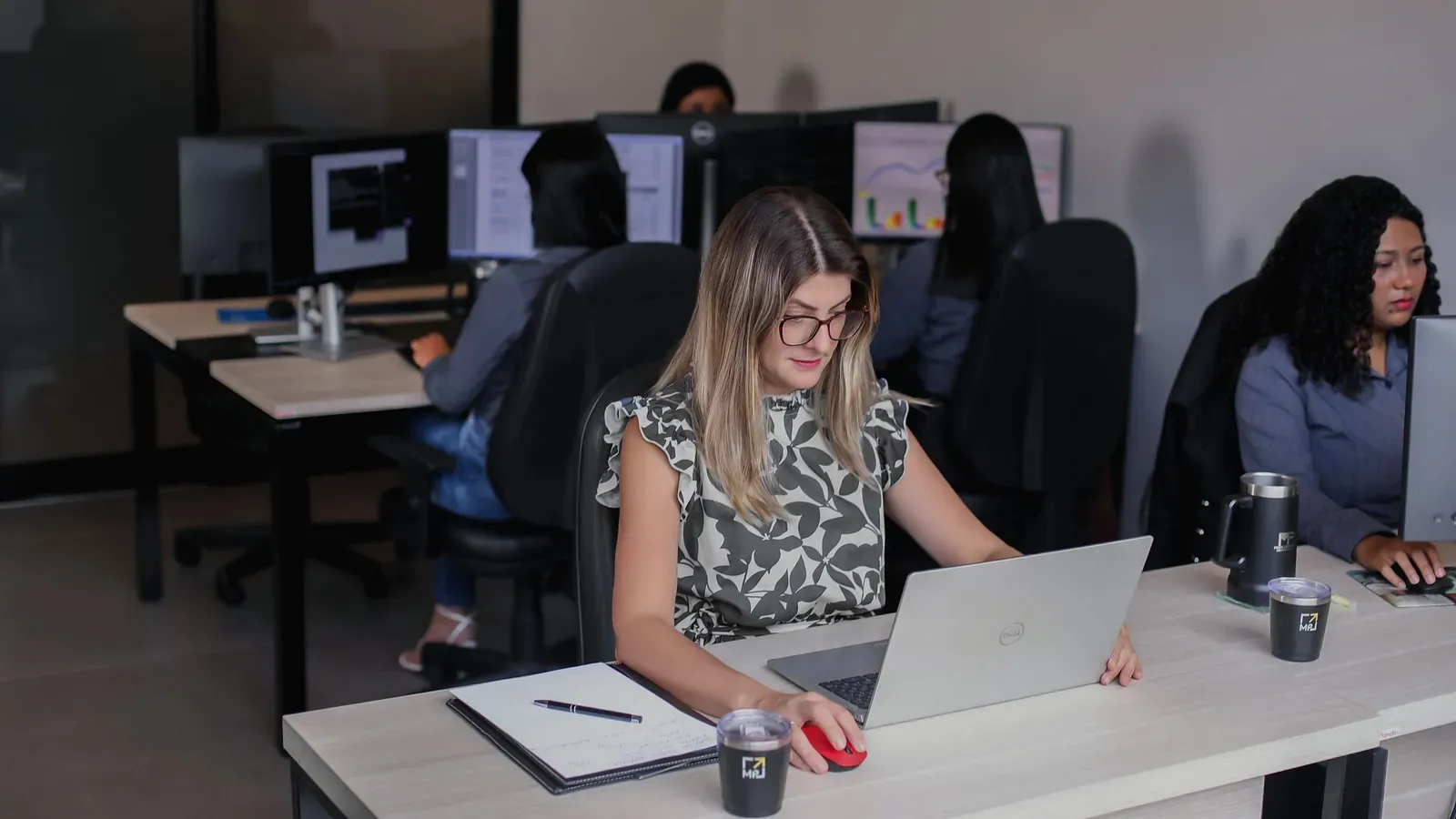 Colaboradora do escritório ao notebook, com a equipe ao fundo nas estações de trabalho